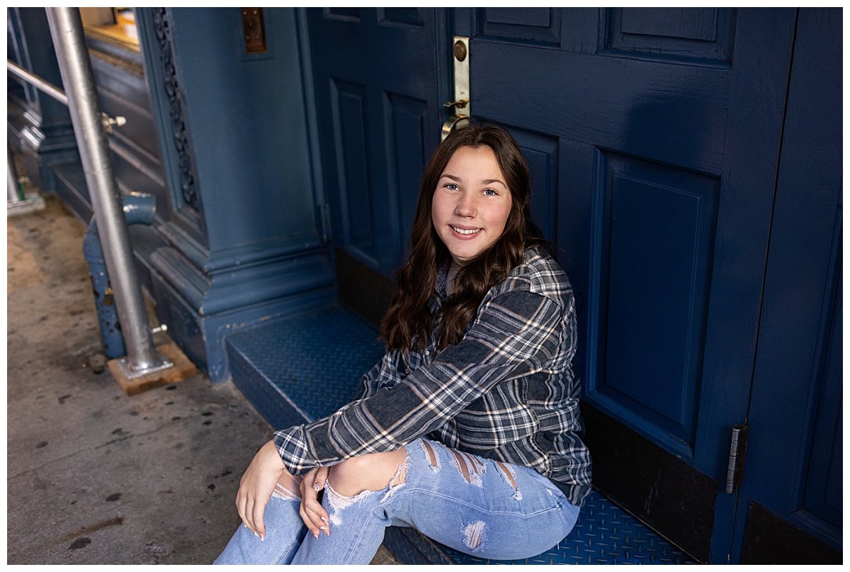 A brunette teen sits next to a blue doorway in NYC during a 16th birthday travel session taken by Kellie Rochelle Photography.