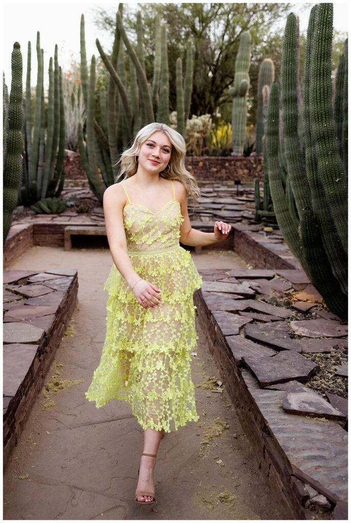 A young woman with blonde hair wearing a yellow lace dress and tan heels walks amongst the cacti during her Arizona travel senior session.