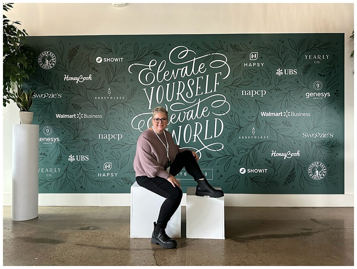 A blonde photographer in a mauve sweater and black leggings poses in front of the Elevate Creative Summit backdrop in Nashville, TN.