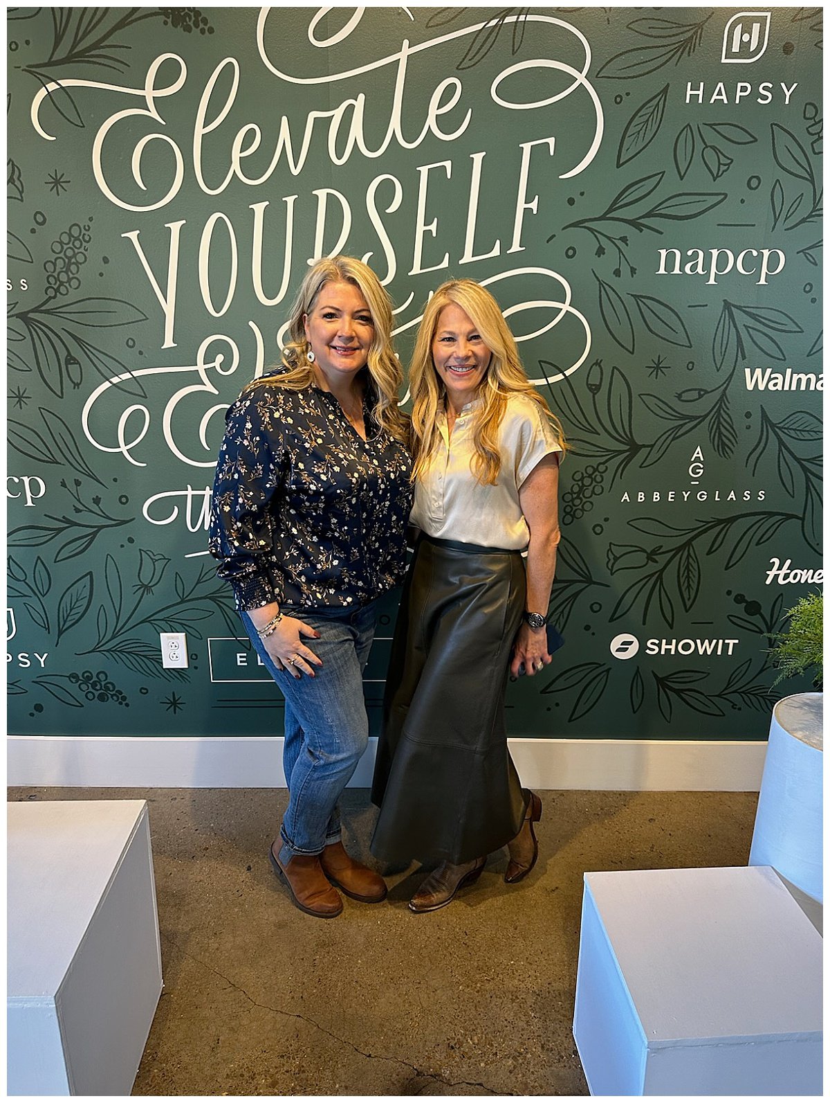 Two blonde women pose for a photo in front of a green sign that reads in white script font, "Elevate yourself," in Nashville, TN.