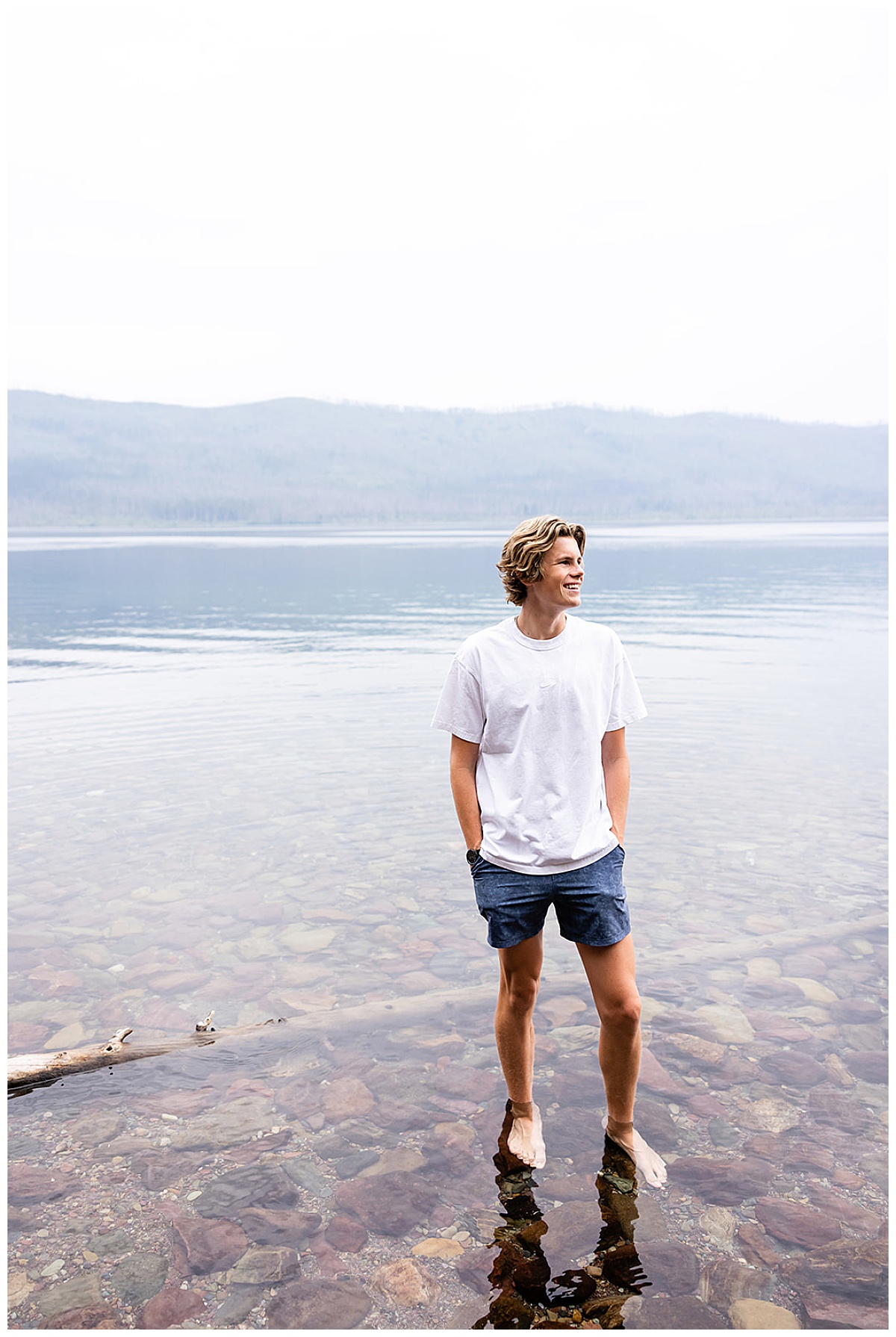 A senior boy wearing a white t and blue shorts stands barefoot in the crystal clear water at Glacier National Park