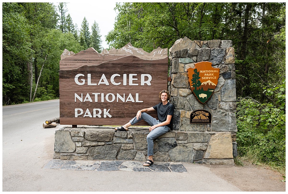 A teen boy wearing a black tshirt and jeans with Birkenstock sandals sits against the stone National Park Service sign for Glacier National Park in Montana.