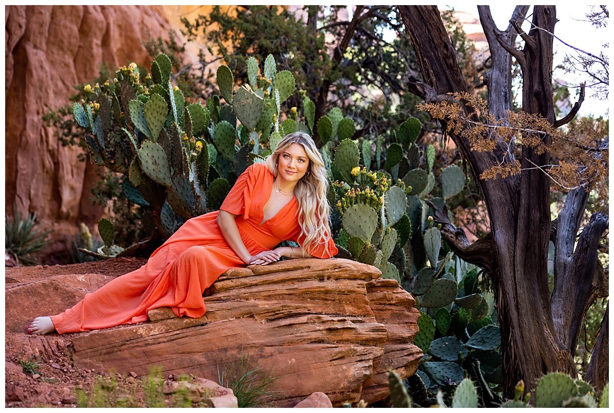 A young woman with long, blonde hair wearing a floor-length orange dress lounges on the red rocks at Fay Canyon Overlook in front of cacti during a travel photo session in Sedona, Arizona.