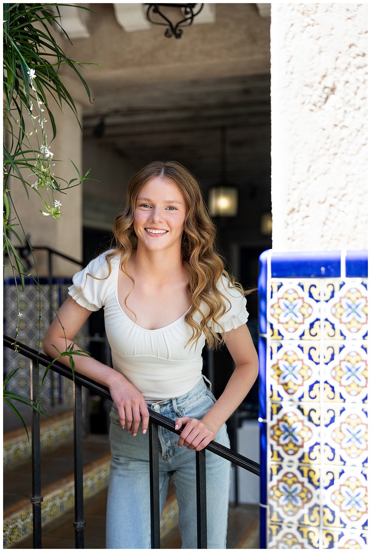 Wearing a white cap sleeve blouse and light wash jeans, a high school senior leans over a black iron handrail next to blue, orange, and white southwestern style tiles during an Arizona senior session.