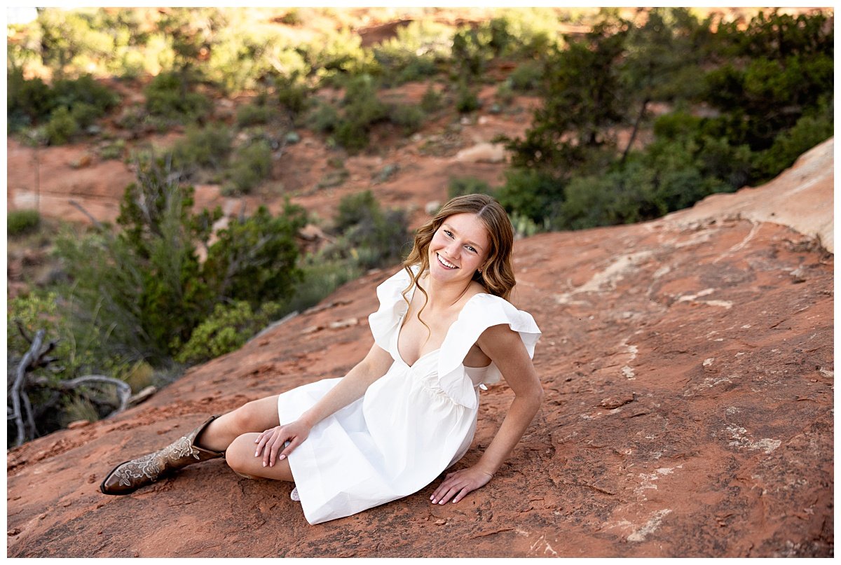 A Williston, ND high school senior is photographed in a white dress and brown cowboy boots sitting atop a red rock formation at Fay Canyon Overlook in Arizona during her travel senior session with Kellie Rochelle Photography.
