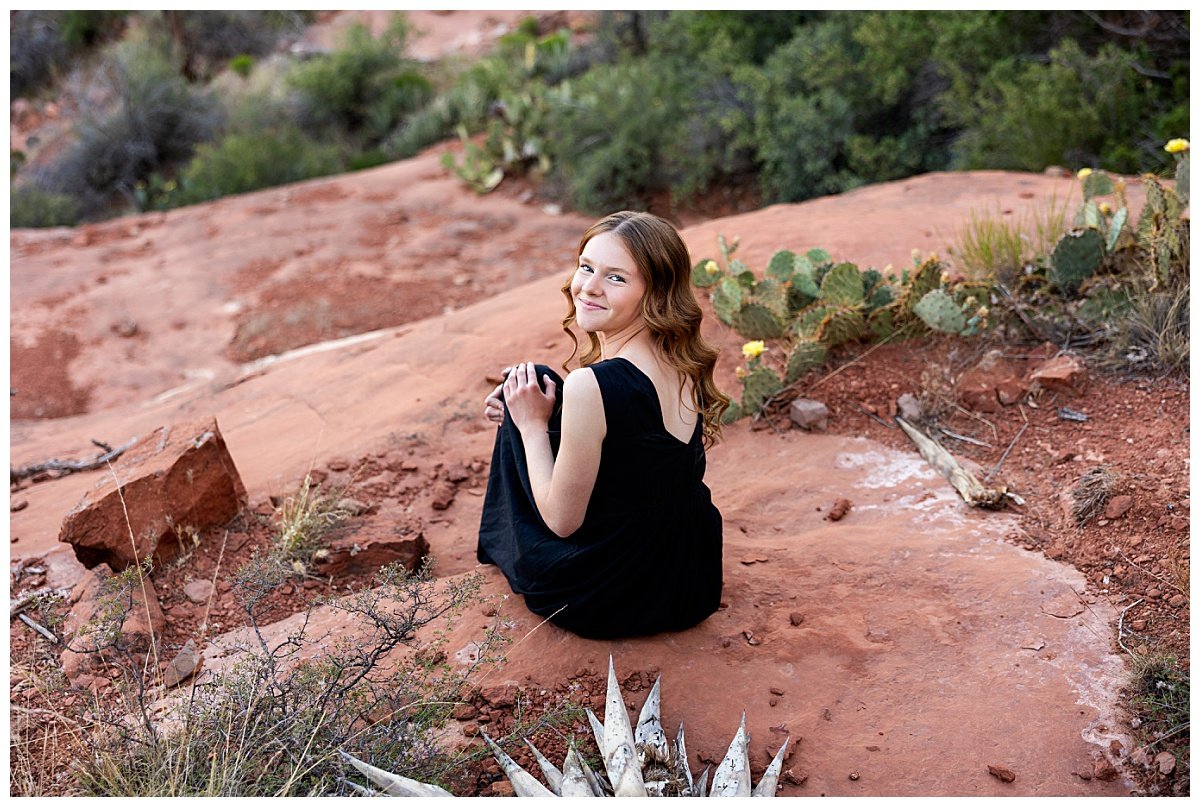 Wearing a black dress and curls in her brown hair, a high school senior is seated upon a red rock formation in Sedona, Arizona, next to cacti during a travel senior session with ND based photographer, Kellie Rochelle Photography.