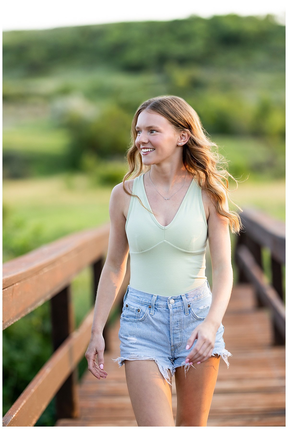 A senior girl with long, brown waves smiles away from the camera wearing a light green halter bodysuit and high-waisted cutoff shorts on a wooden bridge in Lewis and Clark State Park.