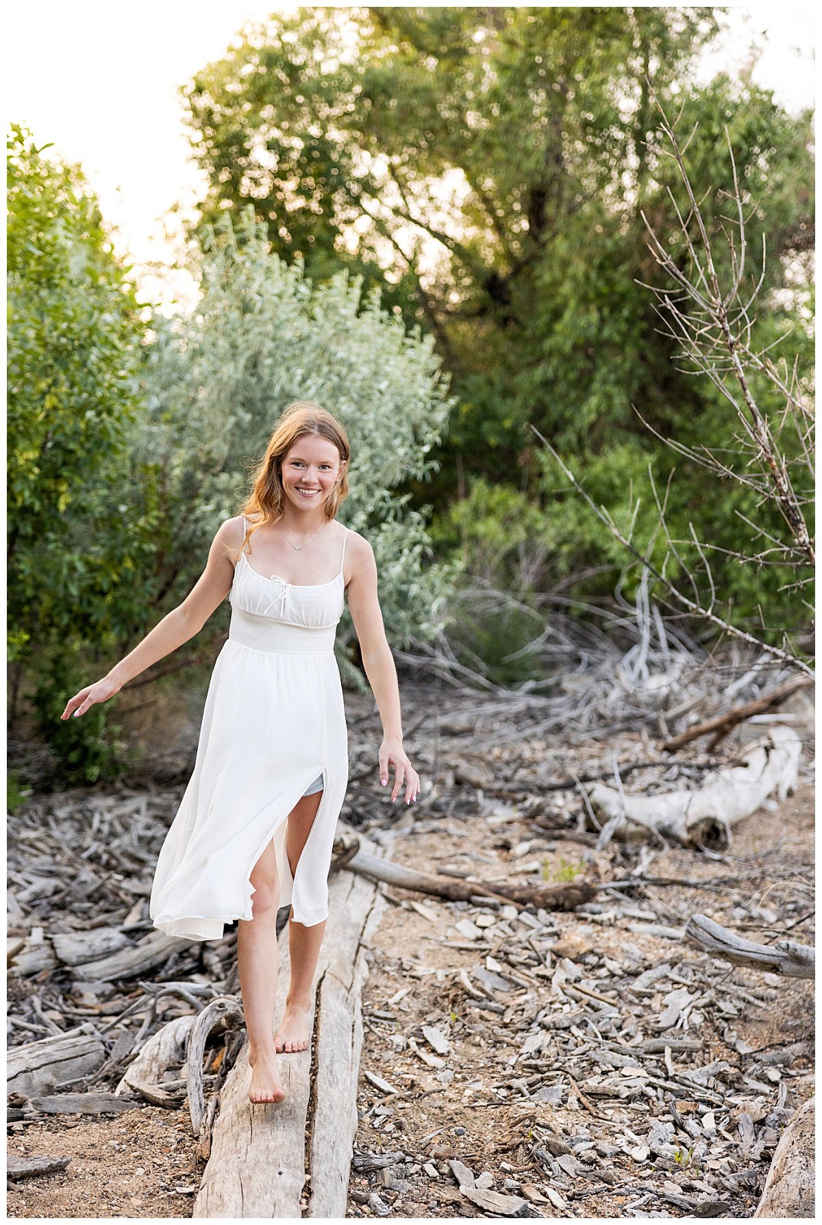 A high school senior is shown balancing on a fallen tree, barefoot in a white dress in Lewis & Clark State Park during her senior photo session with Kellie Rochelle Photography.