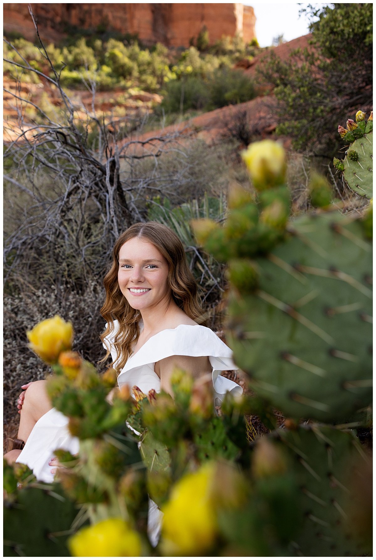 A smiling North Dakota high school senior is seated amongst flowering cacti in the Arizona desert wearing a white dress and boots during a senior photo session with Williston based senior photographer, Kellie Rochelle Photography.