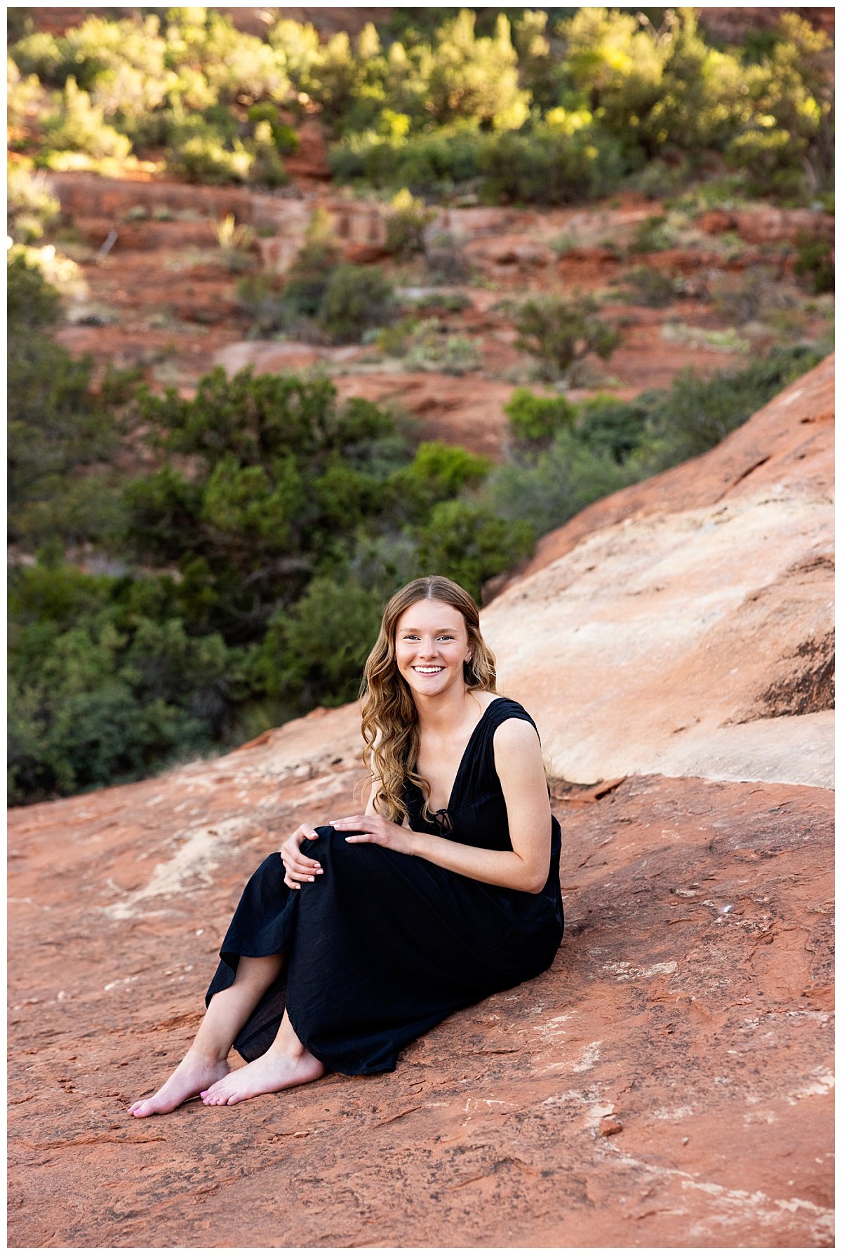 North Dakota based travel photographer, Kellie Rochelle Photography, captures a smiling high school senior with long, brown hair wearing a black, maxi dress seated on an overlook in Sedona, AZ.