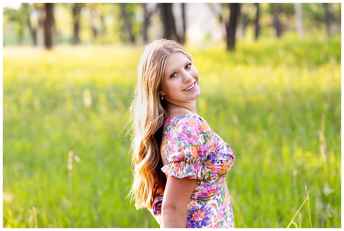 A blonde young woman in a colorful, floral dress looks back over her shoulder at a golden hour senior session with North Dakota based senior photographer, Kellie Rochelle Photography.