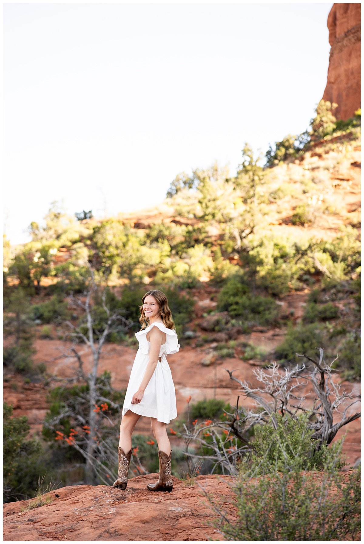 A brunette high school senior smiles over her shoulder in a white knee-lenght dress and brown cowboy boots at the base of butte in Sedona, Arizona, during her shoot with North Dakota based travel photographer, Kellie Rochelle Photography.
