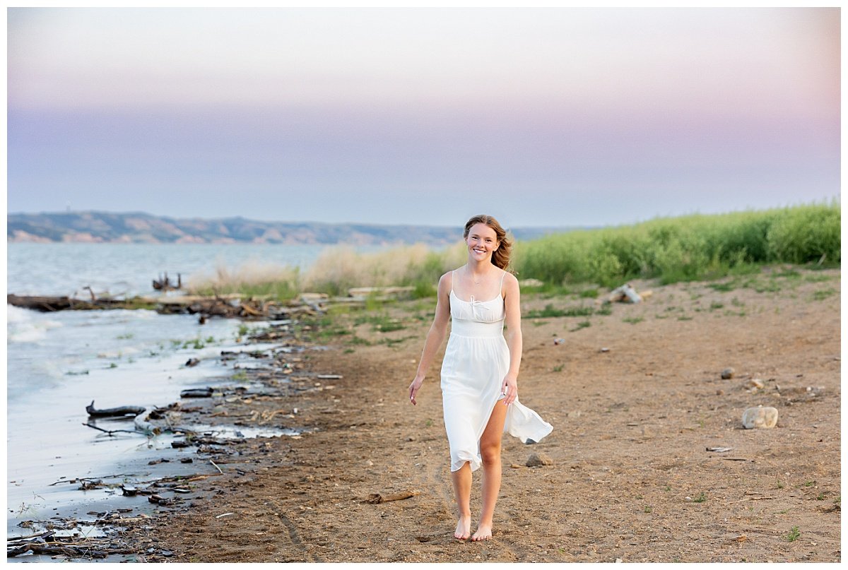 A brunette high school senior strolls next to a lake at Lewis and Clark State Park set against a lavender and blue sunset in the background.