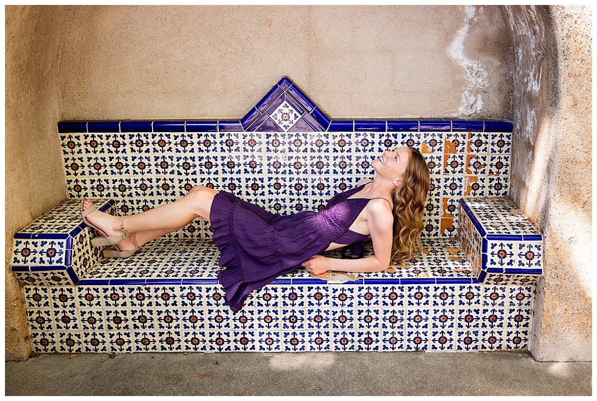 A brunette senior girl wearing a deep purple dress and tan block heels lounges on a southwestern tiled bench comprised of deep blue and orange designs during her senior session in Sedona, AZ.