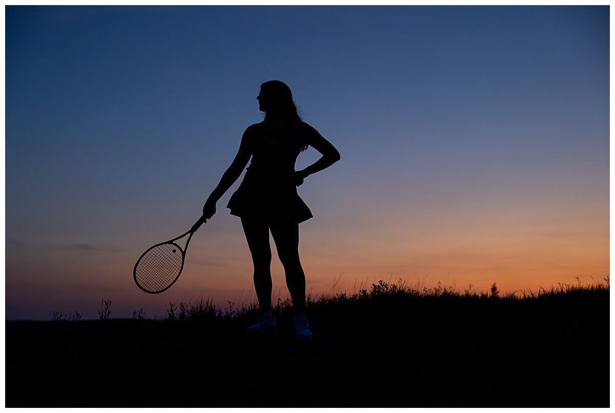 A silhouette of a high school senior holding a tennis racket is shown before an orange an deep blue sunset at blue hour in Theodore Roosevelt National Park.
