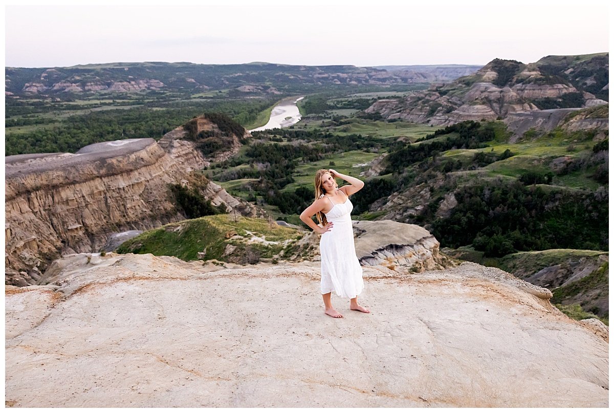 A senior girl is photographed wearing a white dress on a cliff side in North Dakota with a scenic view of Theodore National Park in the background.