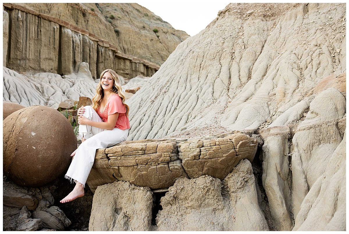 A young woman smiles brightly in her white pants and coral top holding her knee to her chest during a North Dakota senior photoshoot with Kellie Rochelle Photography.