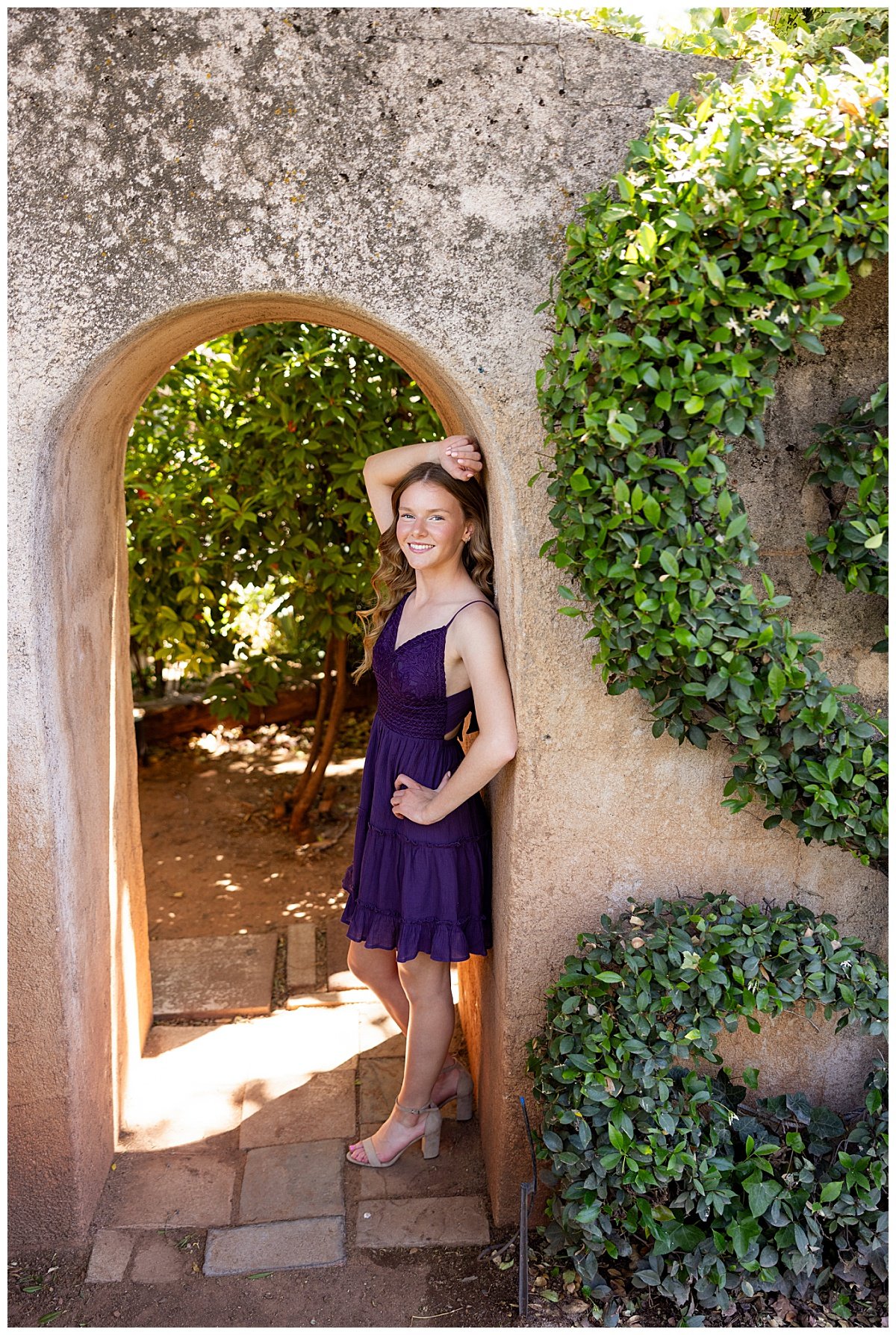 A senior girl wearing a knee-length, deep-purple dress leans against a stucco archway with one arm lifted above her head while her other hand is placed on her hip during a senior session in Arizona.