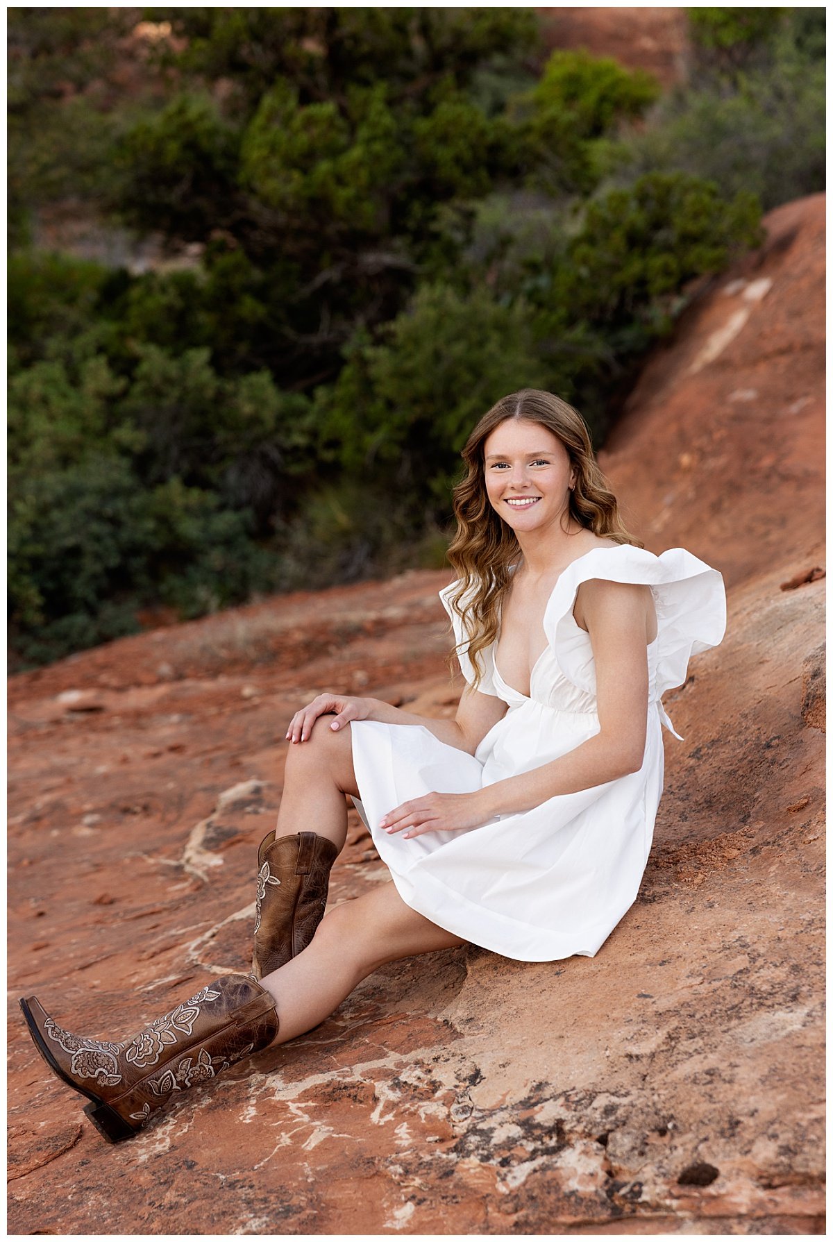 A Williston, ND high school senior is photographed in a white dress and brown cowboy boots sitting atop a red rock formation at Fay Canyon Overlook in Arizona during her travel senior session with Kellie Rochelle Photography.