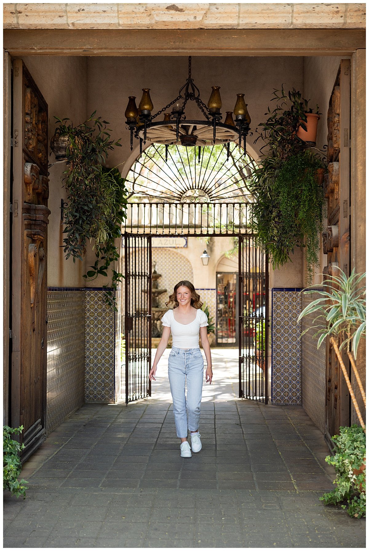 Opting for a more casual wardrobe for her senior photo session with Kellie Rochelle Photography, a North Dakota high school senior walks under a wrought iron chandelier in an Arizona breezeway wearing a white cap-sleeved blouse.
