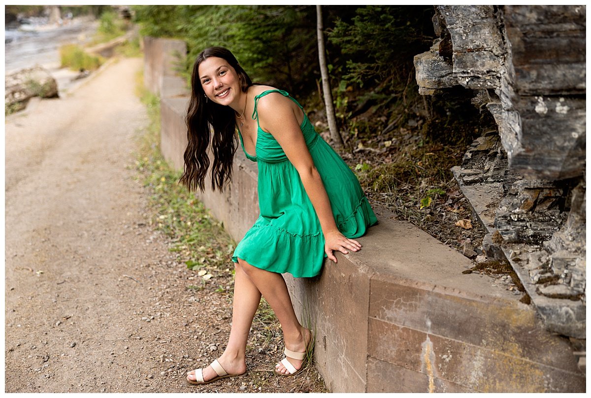A brunette high school senior wearing a knee-length green dress and beige sandals is seated on a concrete barrier at Glacier National Park during her senior session with Kellie Rochelle Photography.
