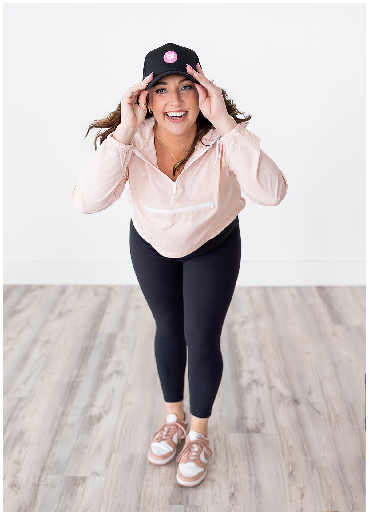 Dark haired beauty influencer, Tara Geltel, smiles before a bright white background wearing black leggings, a pastel pink pull over, and a baseball cap during a branding photoshoot with Williston, North Dakota brand photographer, Kellie Rochelle Photography.