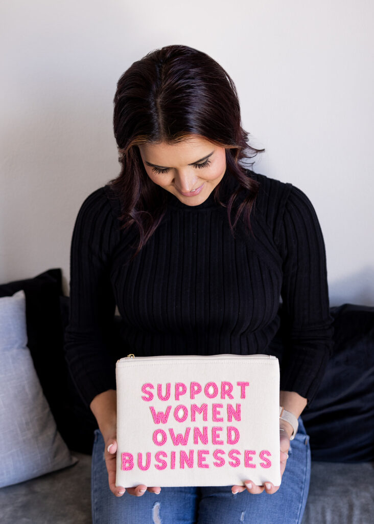 Shandi, owner of North Dakota salon, Creative Elements, is pictured wearing a black sweater, smiling down at a wristlet that reads 'support women owned businesses'.
