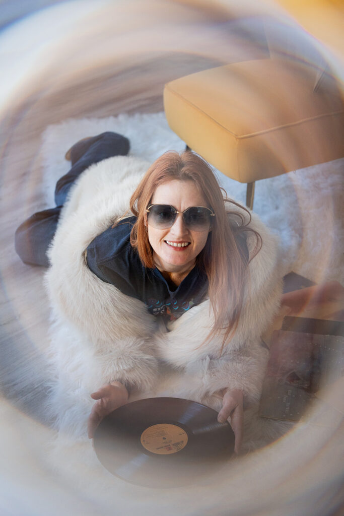 A Williston, ND, woman wearing a fur coat and large 70s aesthetic sunglasses is pictured holding a vinyl album. 