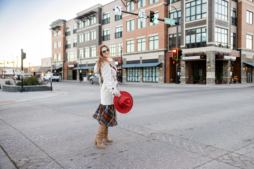 An image from a 70s inspired photoshoot with Williston, ND based photographer is shown.  A woman wearing a plaid skirt and knee high boots holds a burnt orange hat as she crosses an intersection in downtown Williston.