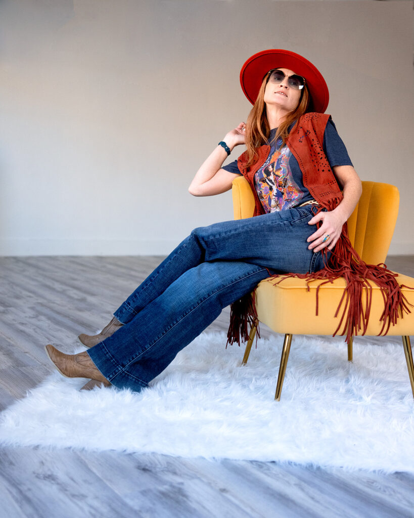A red haired woman wearing bell bottoms, boots, and a burnt orange fringe vest over a band t is seated on a mustard yellow chair in a Williston, ND photo studio for her 70s vintage inspired photoshoot with Kellie Rochelle Photography.