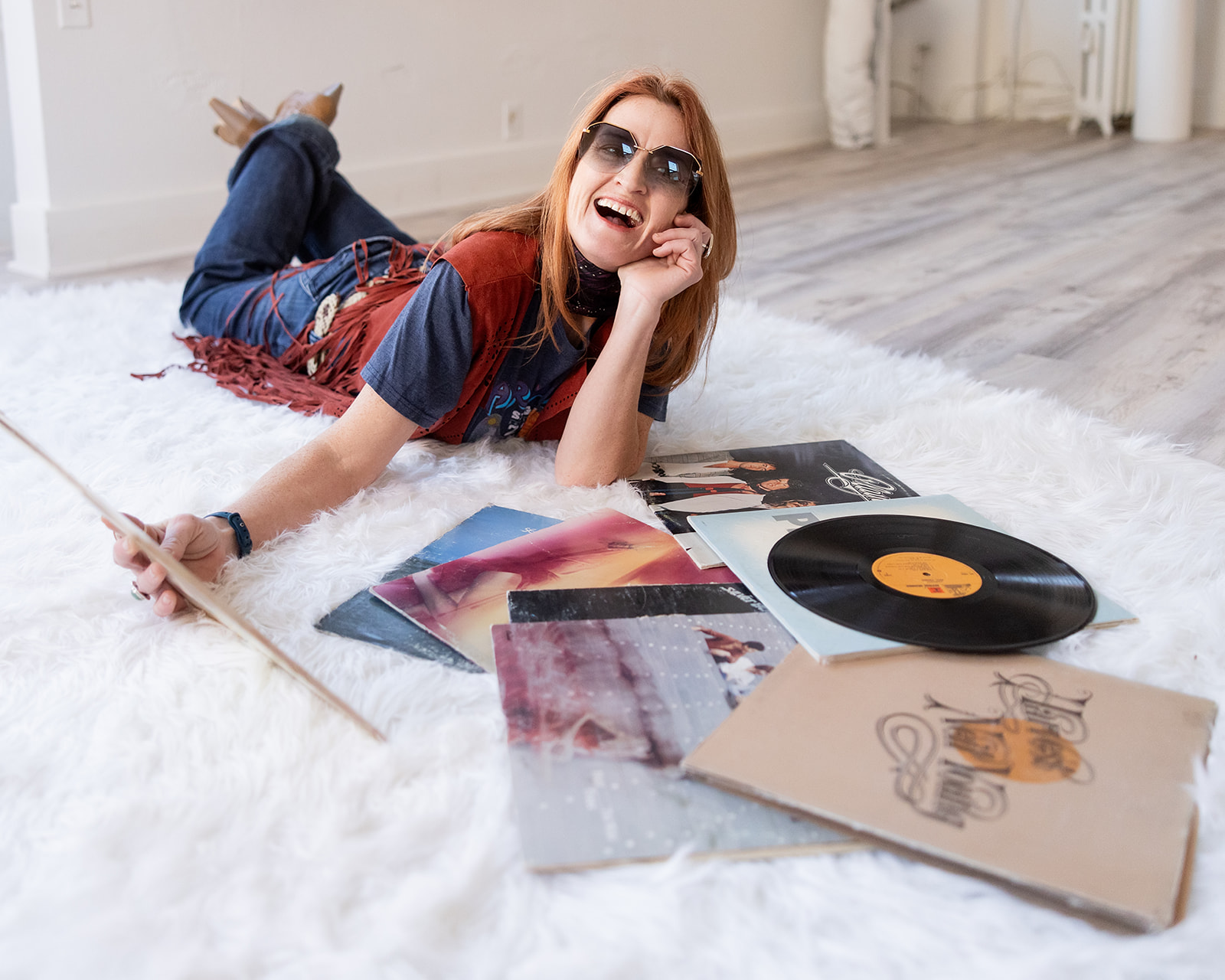 A red haired woman wearing sunglasses and a 70s inspired wardrobe laughs as she lies on the floor of a Williston, ND photo studio surrounded by vintage vinyls during her 70s vintage inspired photoshoot with Kellie Rochelle Photography.