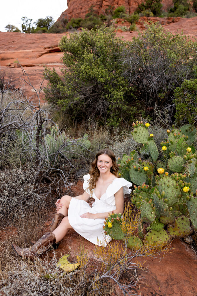 A North Dakota senior  is shown seated next to cacti in Sedona, AZ, during a travel senior photoshoot with Kellie Rochelle Photography.