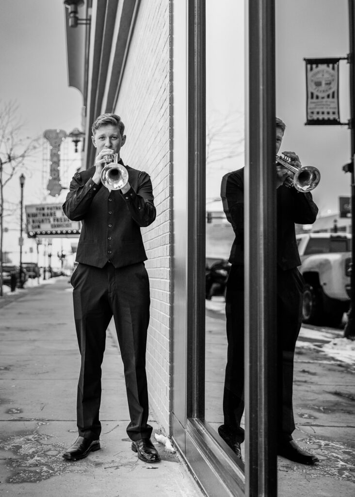 A North Dakota senior boy is shown in a black and white photo from his class of 2024 senior session playing his trumpet in downtown Williston.