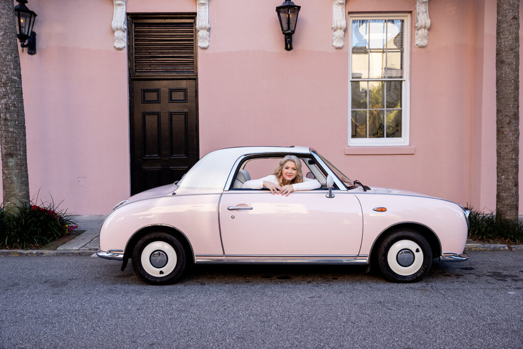 Kellie of Kellie Rochelle Photography is pictured in the front seat of a small pink convertible next to a palm tree in South Carolina during a photography retreat in Charleston. 