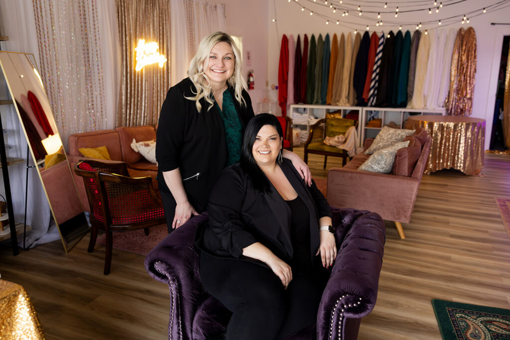 North Dakota event planners, Ashley & Angie, of Williston based business, House of Style, are pictured in the lobby of their building wearing black blazers before a background of fabrics used for event decor. 