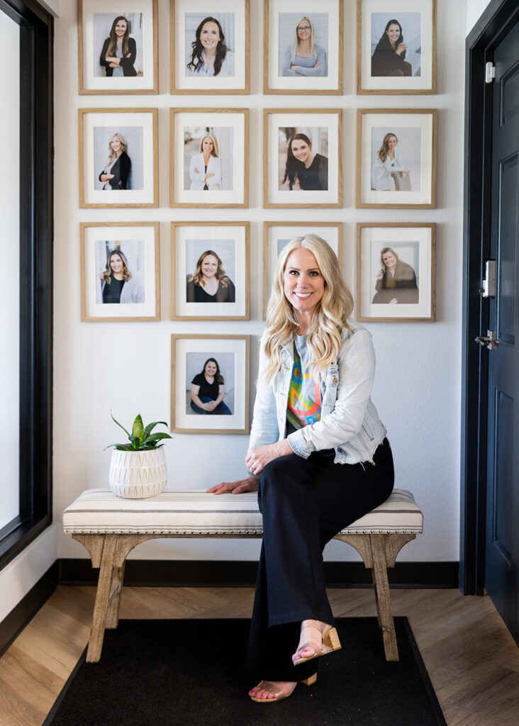 Williston, ND based behavioral health therapist, Katie Shannon, is photographed wearing a jean jacket, heels, and black pants, seated before a gallery wall of the 13 staff members who make up the team at ConnectUs therapy during a woman owned business photoshoot with Williston, ND branding photographer, Kellie Rochelle Photography.