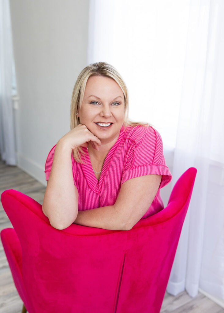 North Dakota woman owned business owner, Angela Skogen, poses in a pink shirt seated on a deeper pink velvet accent chair in the studio of Williston, ND photographer, Kellie Rochelle Photography.