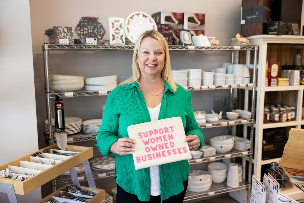 Angela Skogen of Cooks on Main in Williston, ND holds a pouch that reads, "SUPPORT WOMEN OWNED BUSINESSES," as she stands before a selection of plates & bowls available in her storefront during a woman owned business branding session.