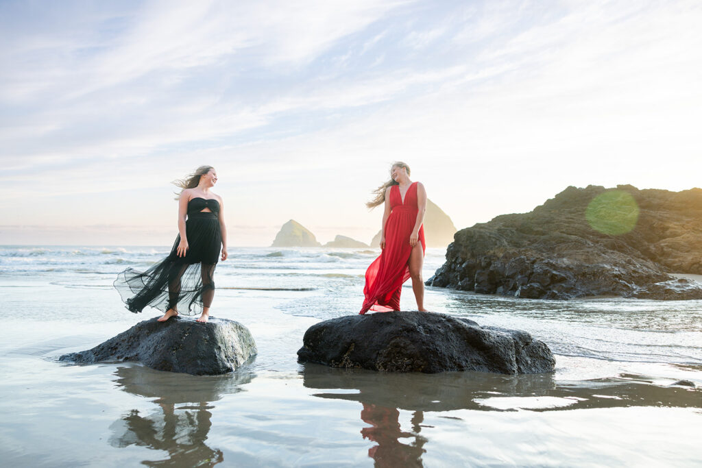 Two senior girls wearing flowing dresses, one black & one red, stand atop rocks exposed at the Pacific shoreline at Short Beach during an Oregon senior session with Kellie Rochelle Photography.