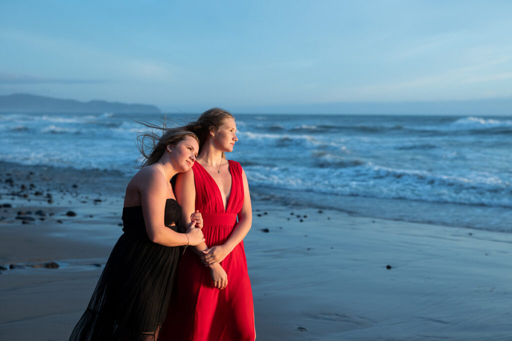 Two senior girls wearing flowing dresses, one black & one red, look out towards the Pacific ocean as waves crash against the shore at Short Beach during an Oregon senior session with Kellie Rochelle Photography.