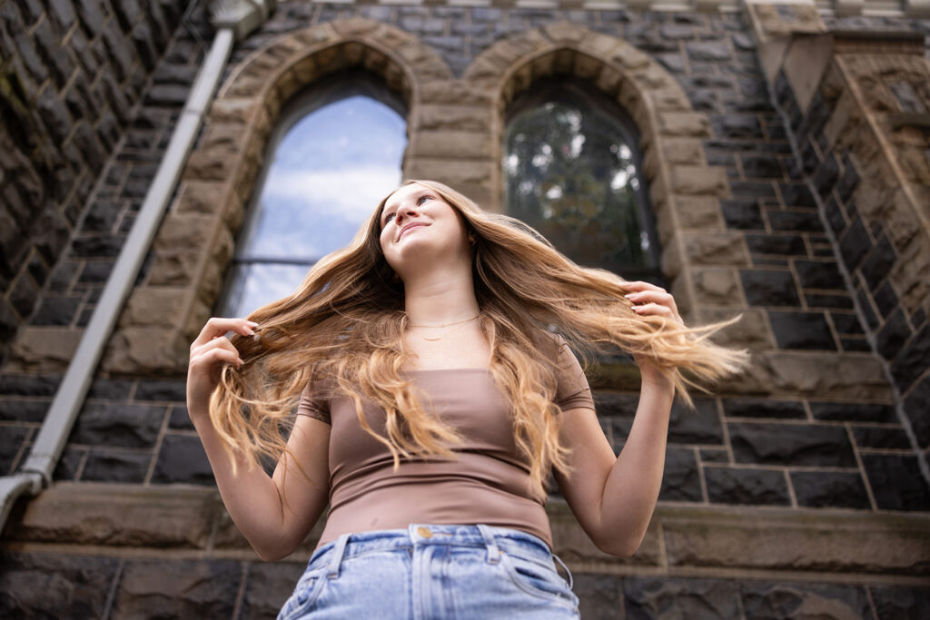 A blonde young woman wearing jeans and a brown square neck top stands before a brownstone building during her downtown Portland senior photoshoot.