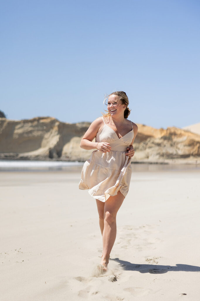 A North Dakota senior girl wearing a cream colored spaghetti strap dress runs, laughing along the Pacific shoreline at Cape Kiwanda during an Oregon Senior session with Kellie Rochelle Photography.