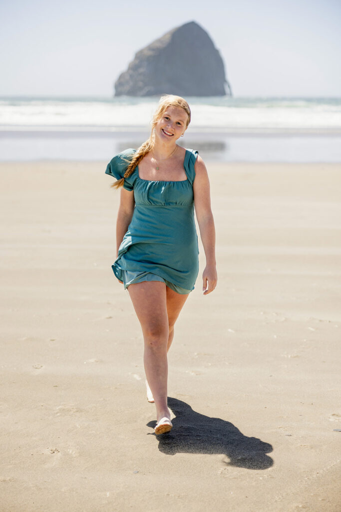 A North Dakota senior girl wearing a teal cap sleeve dress walks away from the Pacific shoreline at Cape Kiwanda during an Oregon Senior session with Kellie Rochelle Photography.