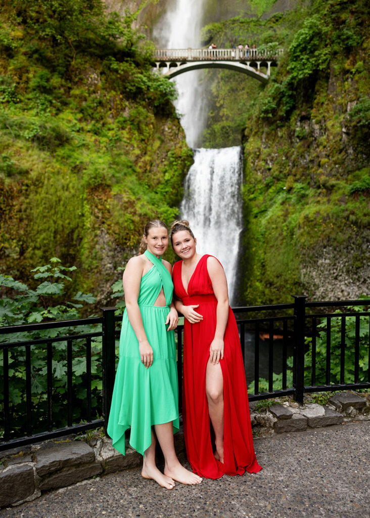 North Dakota senior girls wearing a vibrant green dress and a vibrant red dress lean against a wrought iron railing before an Oregon waterfall surrounded by moss and ferns during an Oregon senior session with Kellie Rochelle Photography.