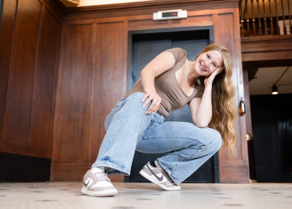 A North Dakota senior with long blonde hair wearing jeans and Nikes crouches before he elevator at the Clyde Hotel during a Portland Oregon senior session with Kellie Rochelle Photography, a North Dakota based travel and senior photographer.