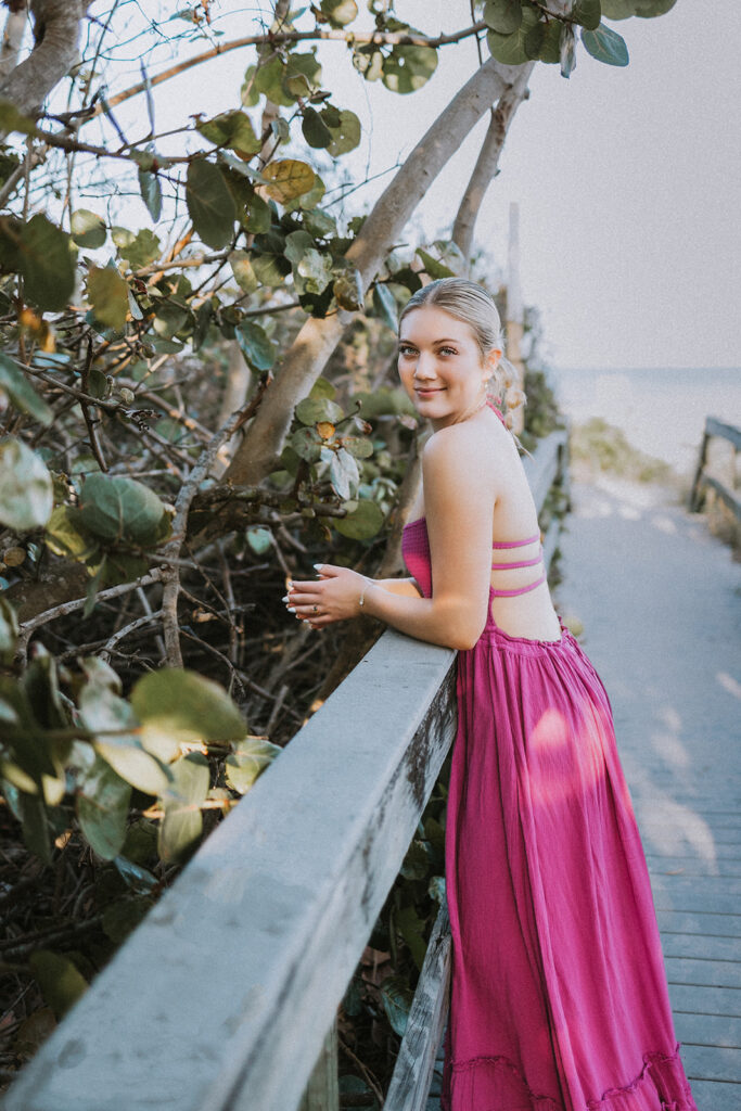 A blonde young woman wearing a long, purple dress leans against the railing of a Florida boardwalk next to the white sand beaches during a travel senior session with Kellie Rochelle Photography.