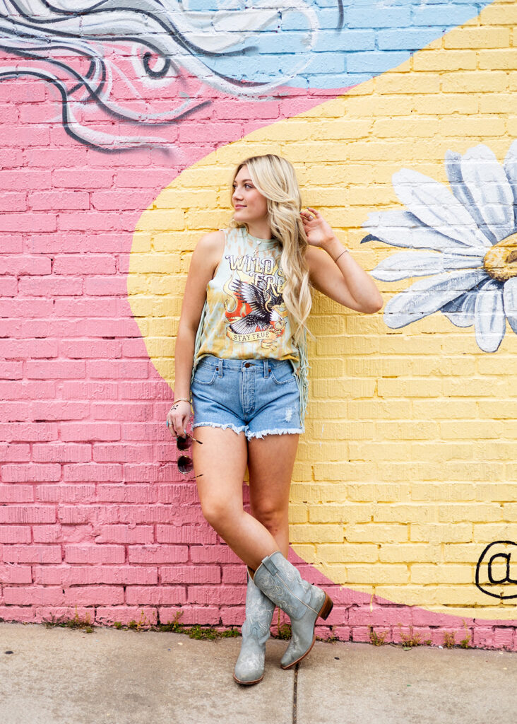 A photo taken by Kellie Rochelle Photography during a travel senior session in Nashville, TN is shown.  A blonde young woman wearing cut offs and boots leans against a colorful painted brick mural.