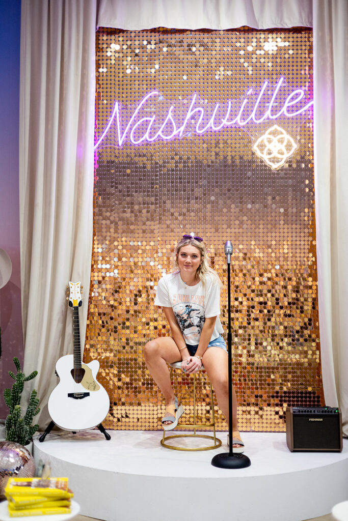 A young woman with blonde hair pinned back with aviator sunglasses is seated on a stool before a gold sequined backdrop with a neon sign that reads "Nashville" on a small stage.  A white acoustic guitar and a small speaker are placed on either side of the Nashville, TN stage.