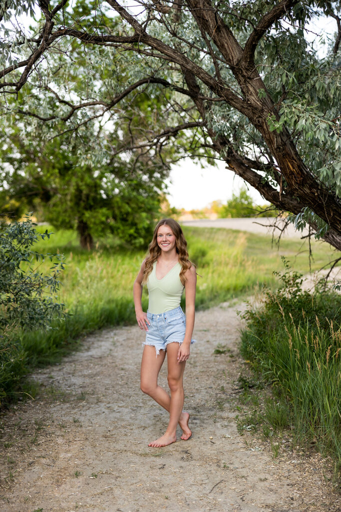 A brunette high school senior wearing a light green body suit and cut off jean shorts is pictured barefoot on a gravel path during a senior photos at Lewis and Clark State Park 