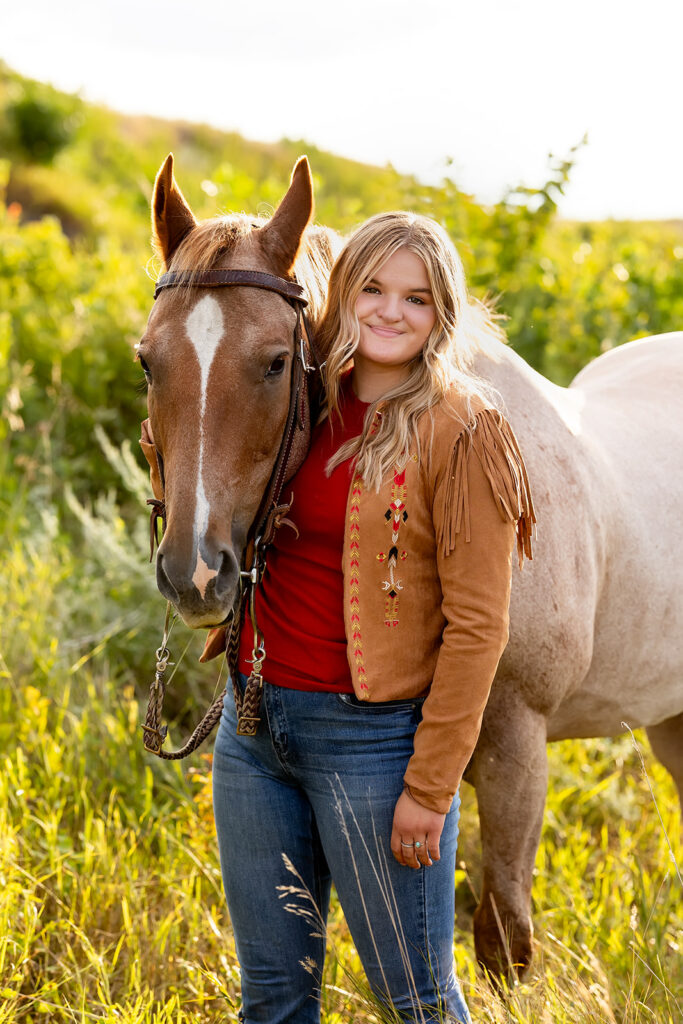 A young woman with blonde hair wearing jeans and a fringed leather jacket sidles next to her brown horse on her family's ranch during her North Dakota senior photos.